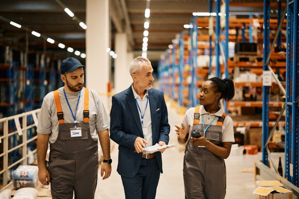 Mature manager communicating with young workers while walking through distribution warehouse.