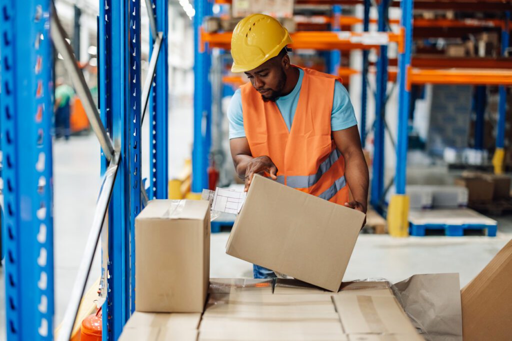 A focused african american warehouse employee carefully lifts a cardboard box in a spacious industrial storage area