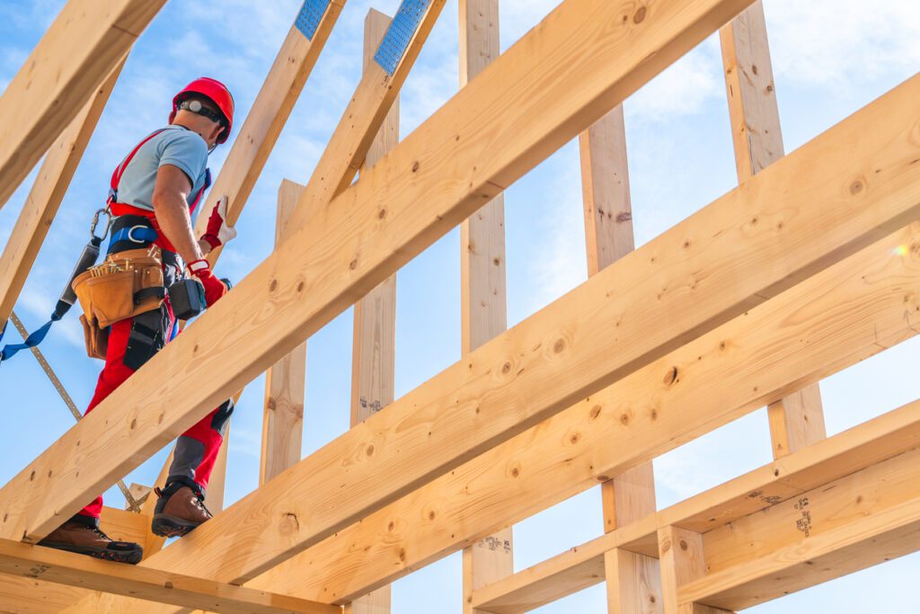 A construction worker wearing a helmet and harness is climbing on wooden beams, working on the roof structure under a clear blue sky in afternoon light.