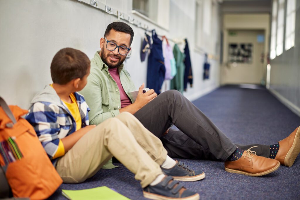 Man working with a child while discussing about learning problem and bullying. Young and friendly teacher speaking to boy in school hallway.