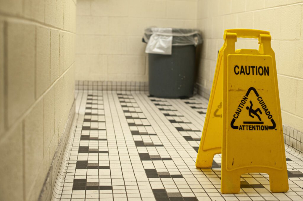 A scuffed caution sign on a black-and-white checkerboard bathroom floor in a public high school. The walls are off-white cinder block, and an industrial gray trash can is visible in the background.