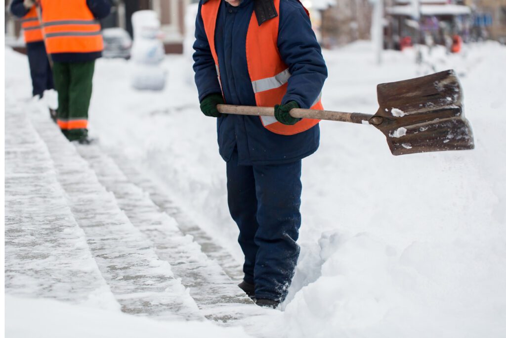 Employees of municipal services in uniforms are clearing snow from the sidewalk with a shovel.