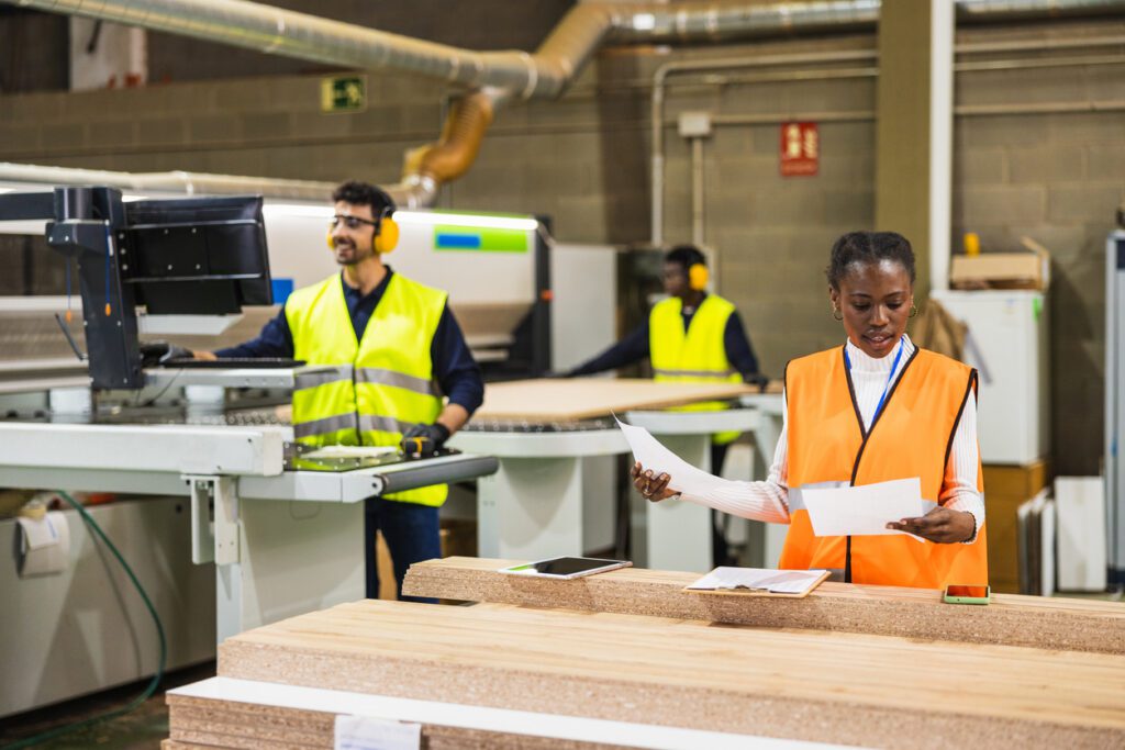 Male and female multi-ethnic workers wearing safety vests inspecting documents and operating machinery in a wood manufacturing factory