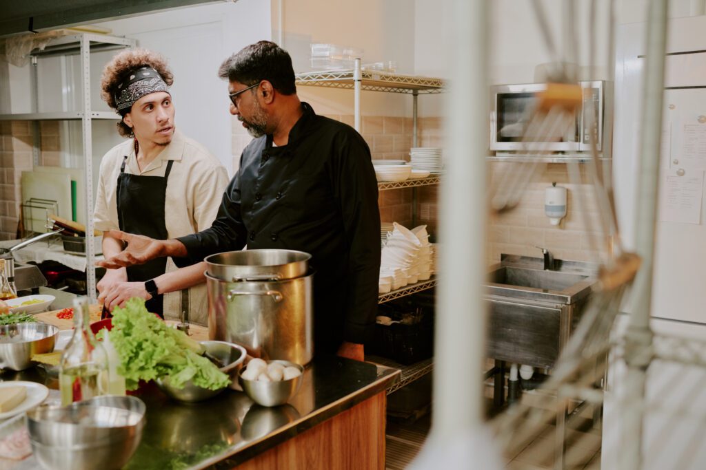 Chef in a black jacket and his sous chef in a white jacket discussing extra ingredients of recipe while sous chef chopping tomatoes