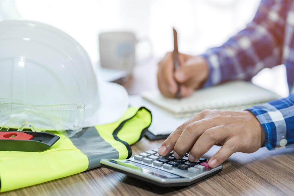 man in construction analyzing revenue calculation recurring income costs using a calculator at a desk with a hard hat and yellow vest