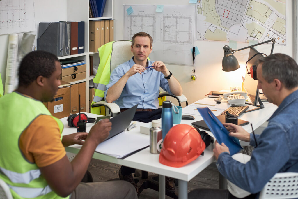 Caucasian middle aged man sitting at desk leading meeting with Black adult man and Caucasian middle aged man in office, discussing construction plans and reviewing documents
